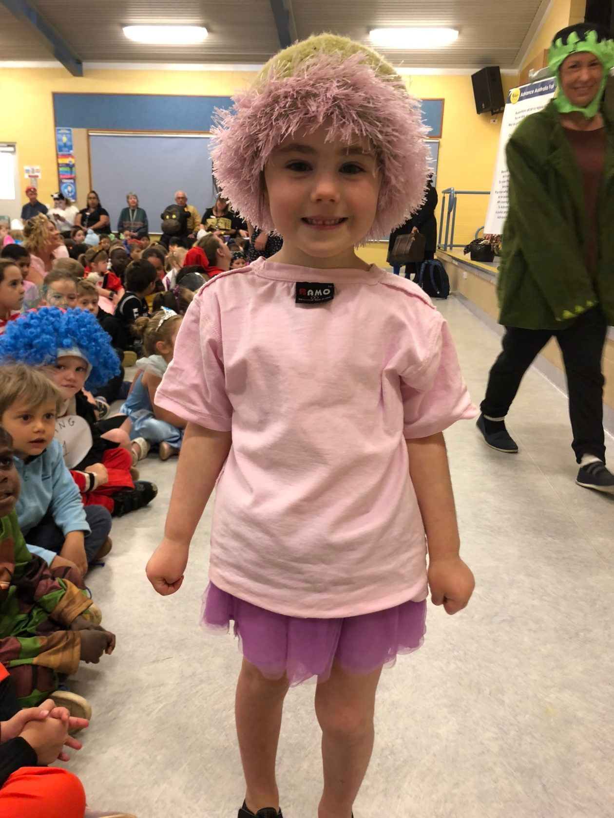 Young girl in pink shirt and purple tutu, wearing a fuzzy hat, smiles at the camera.