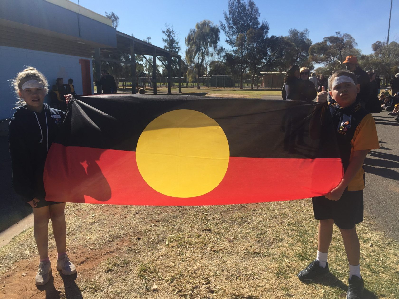 Two kids holding the Aboriginal flag outdoors. They have painted faces, and other people are visible in the background.