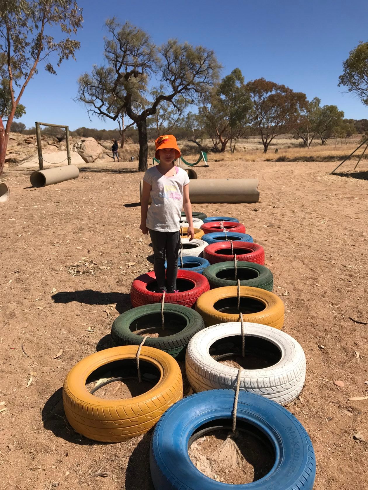 Girl in orange hat walks across colorful tires in a dry, outdoor setting.
