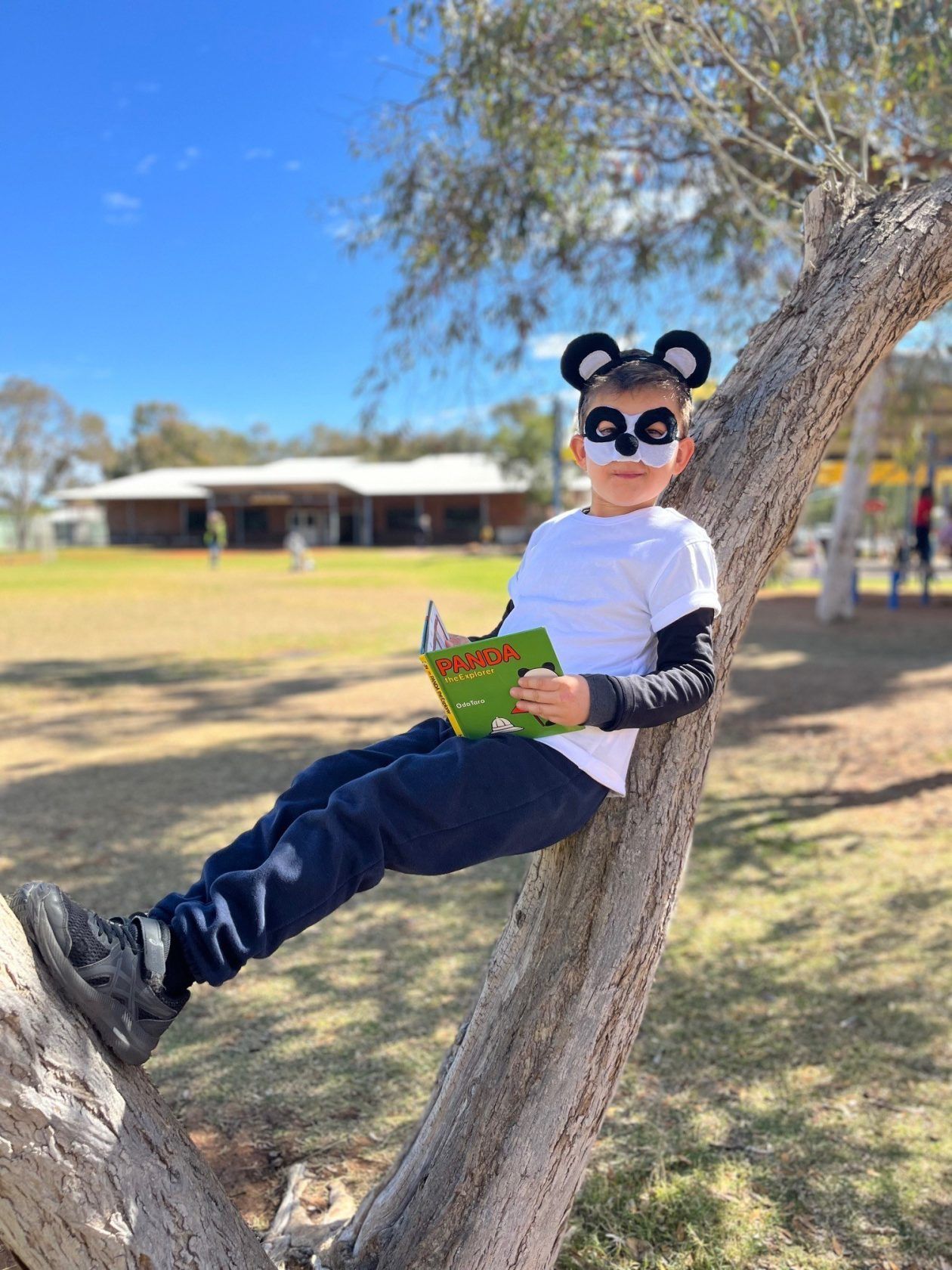 Young boy in panda costume reads a book while sitting on a tree branch outdoors.