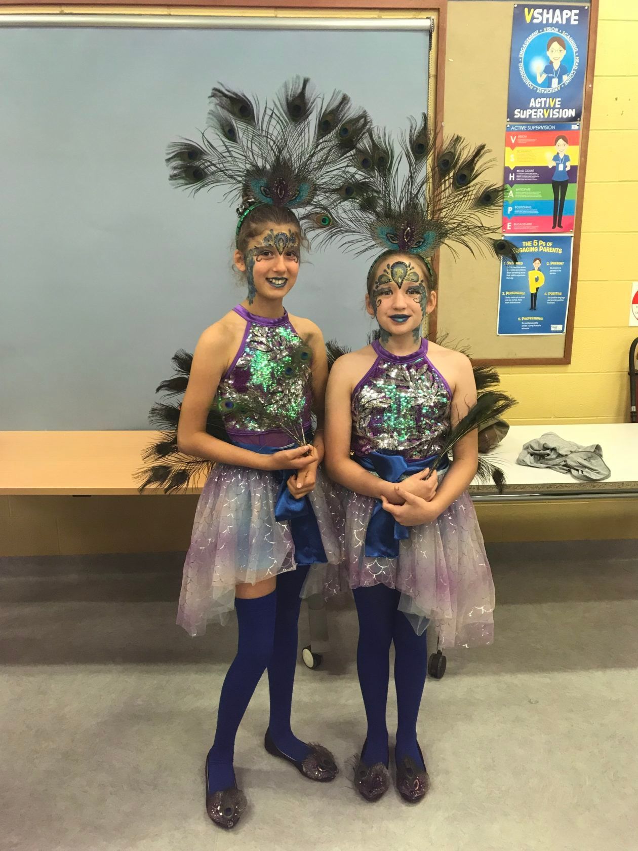 Two girls in peacock costumes smile, wearing feathered headpieces, glittery tops, and blue tights.