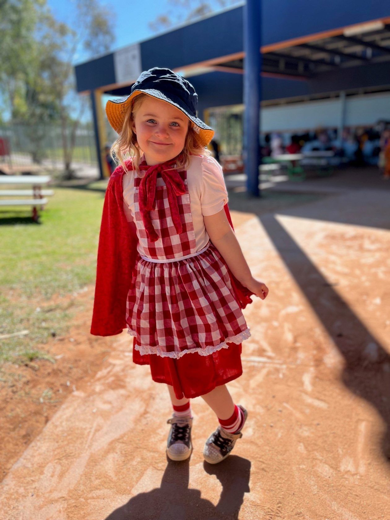 Girl in Red Riding Hood costume smiles outdoors, wearing a red cape, dress, and sun hat.