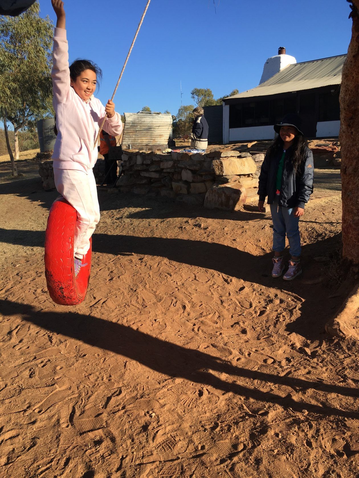 Girl on tire swing, smiling, near a stone building. Another girl watches in the sun. Brown ground.