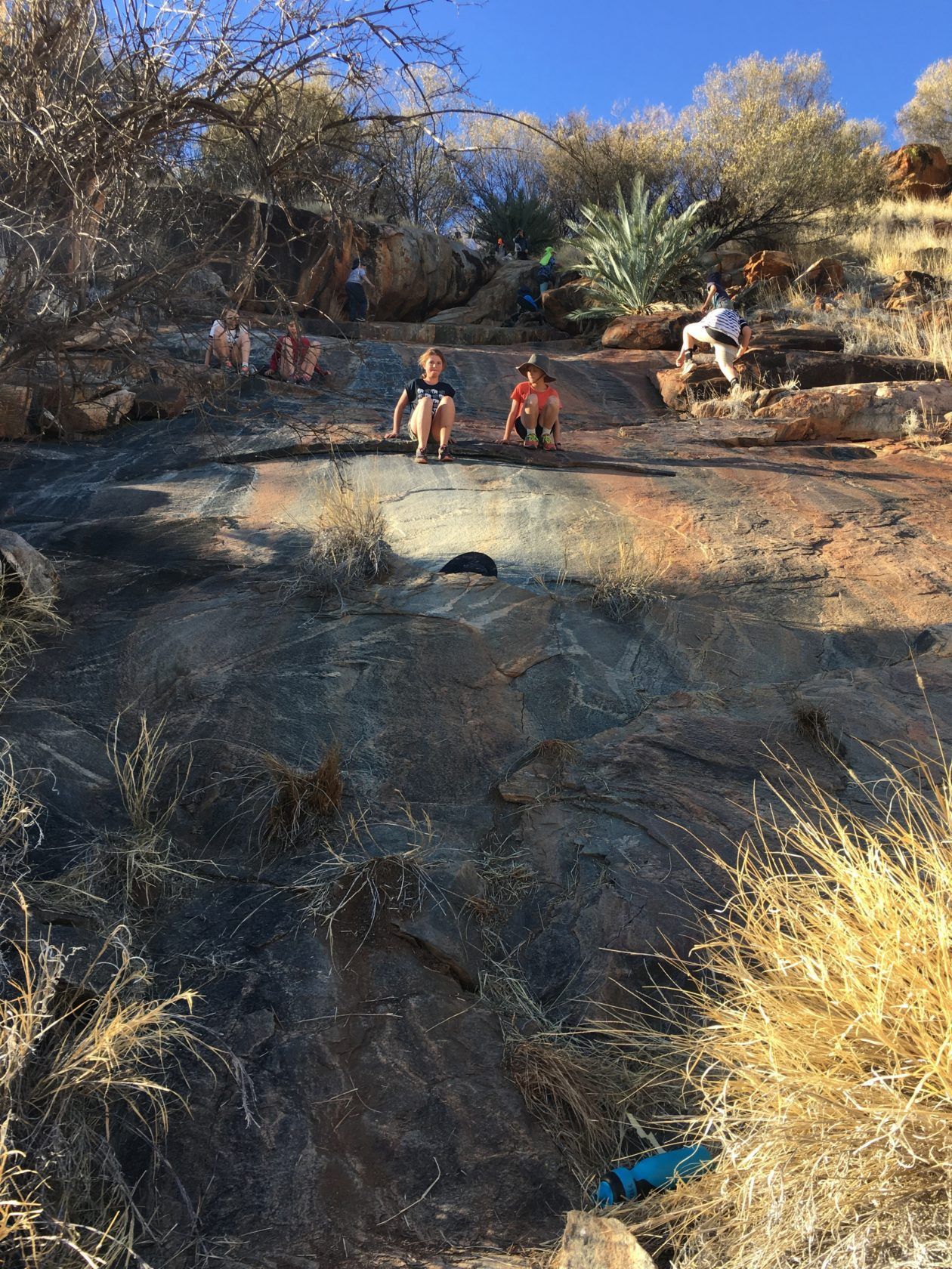 People sliding down a large rock surface in a dry, sunny, outdoor setting.