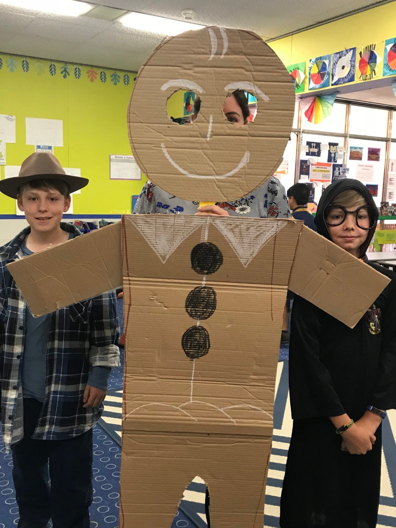 Two children flank a large cardboard gingerbread man, smiling. A person is visible behind the gingerbread man.