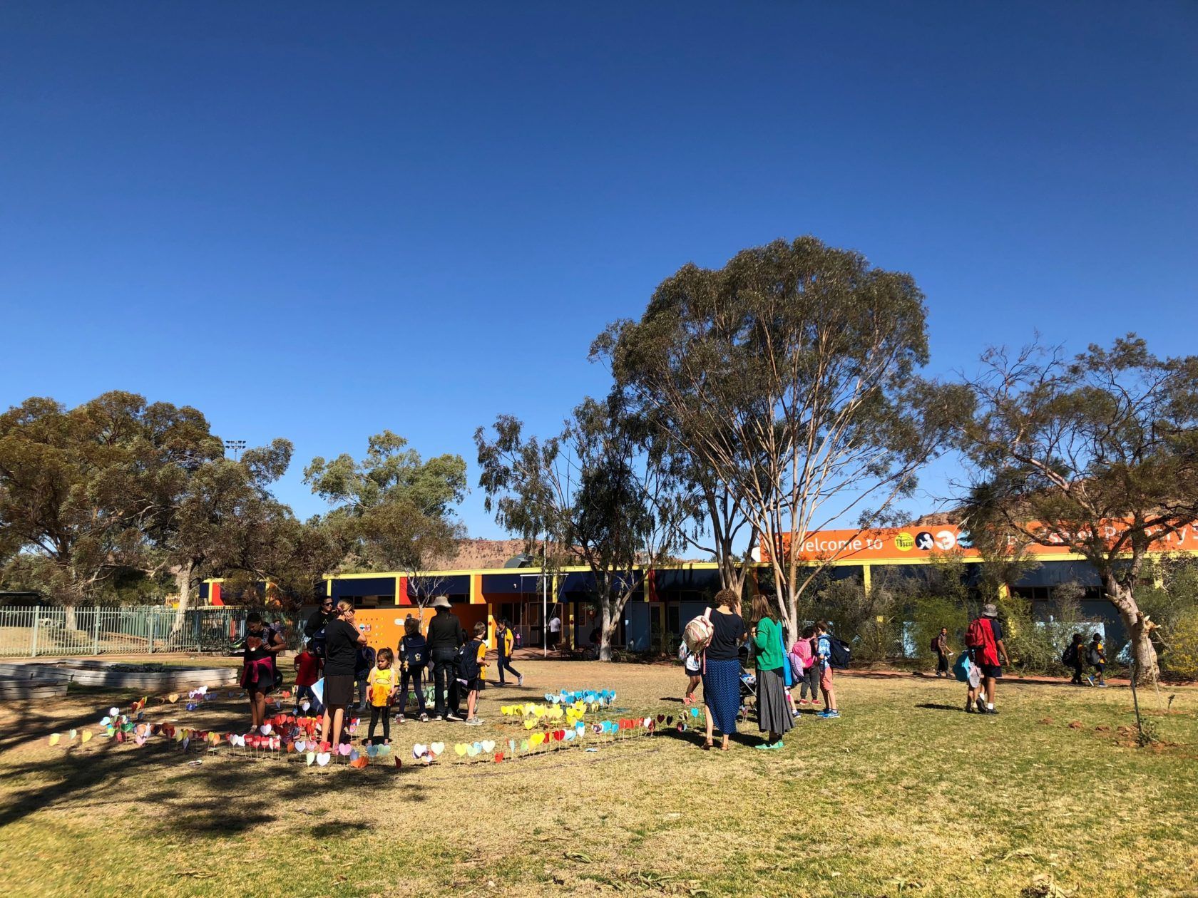 People gather on grass with colorful items spread out; trees and a building in background on a sunny day.