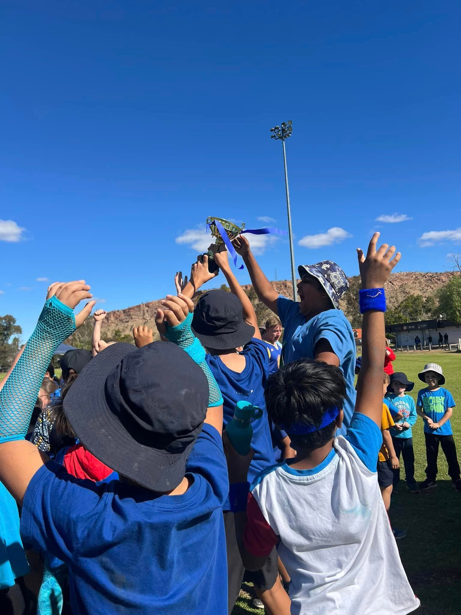 Children in blue celebrate, holding a trophy aloft on a field. Blue sky, sunny day.