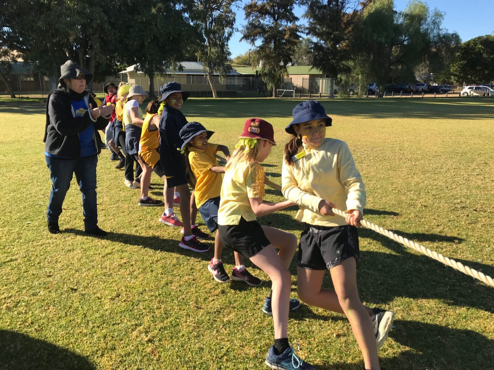 Children in yellow and blue school uniforms playing tug-of-war on a grassy field.