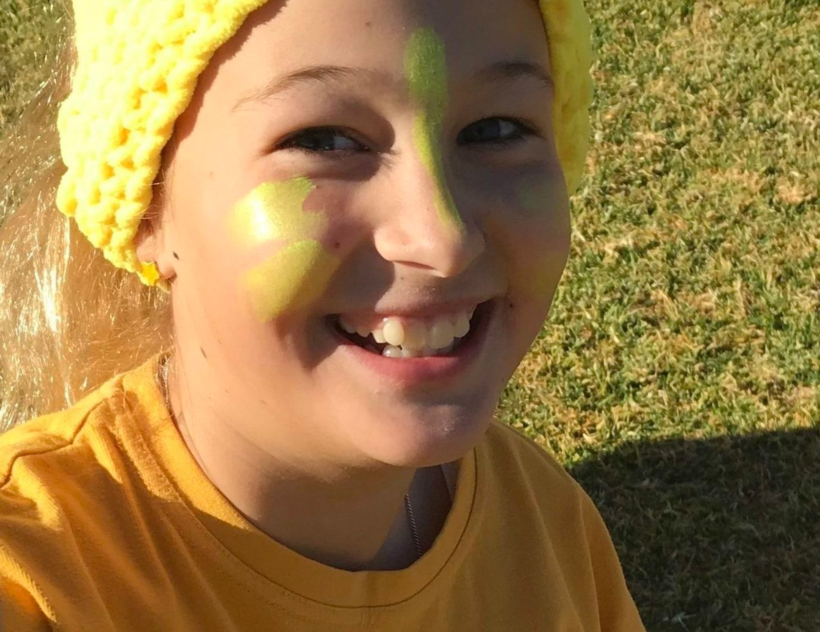 Smiling person with yellow face paint and a yellow hat outdoors on grass.