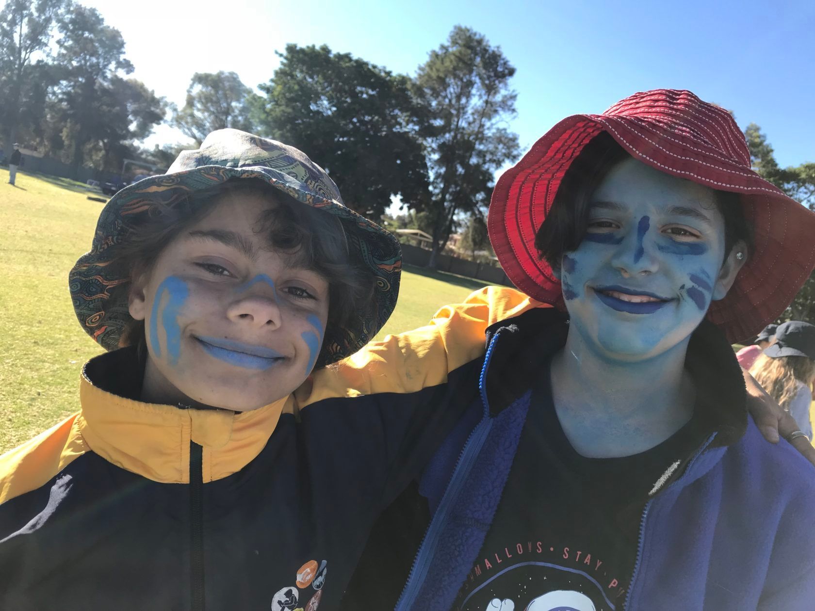 Two smiling kids with blue face paint, wearing hats, outdoors.