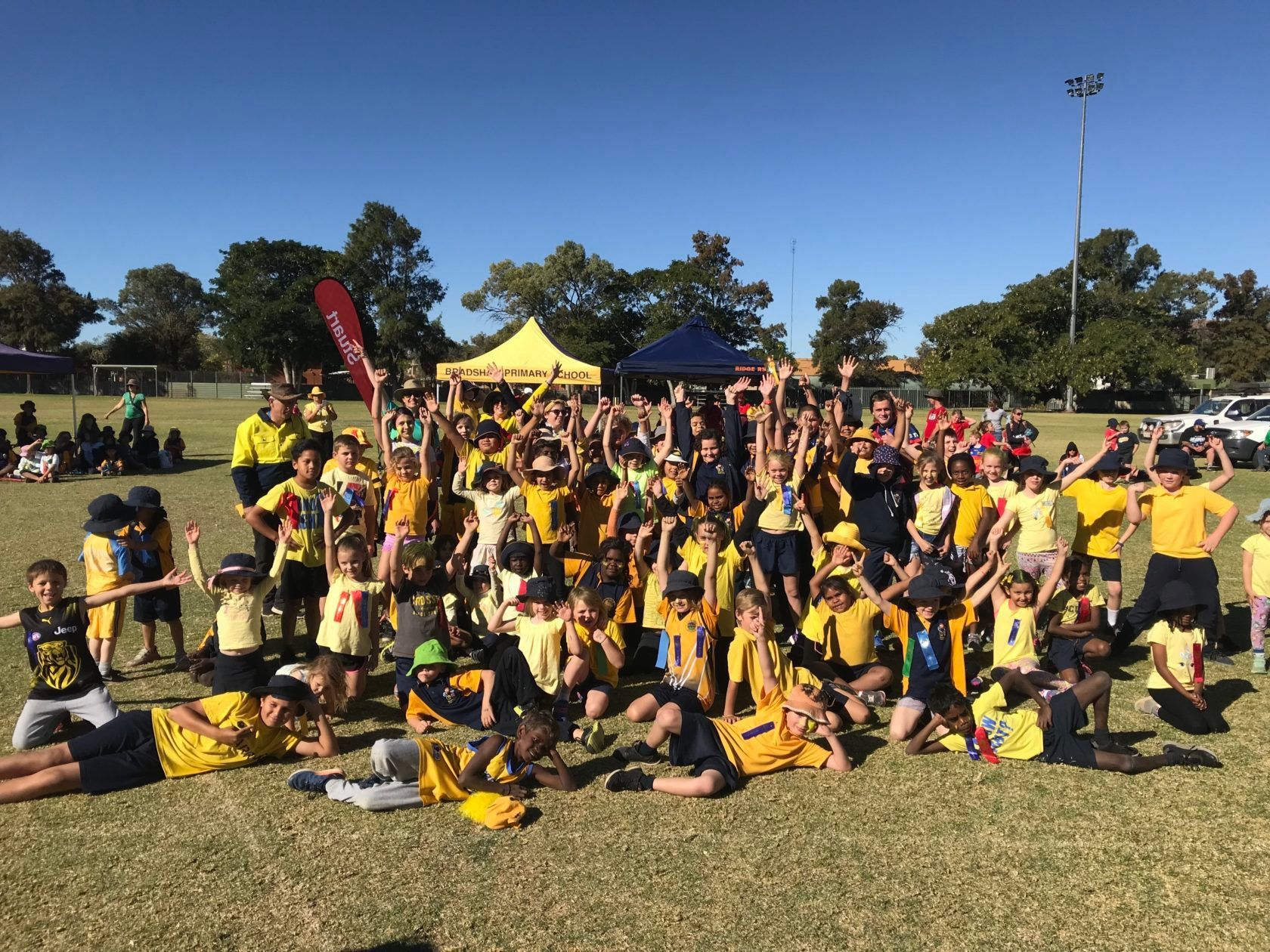 Group of children in yellow shirts pose on a grassy field under a blue sky.