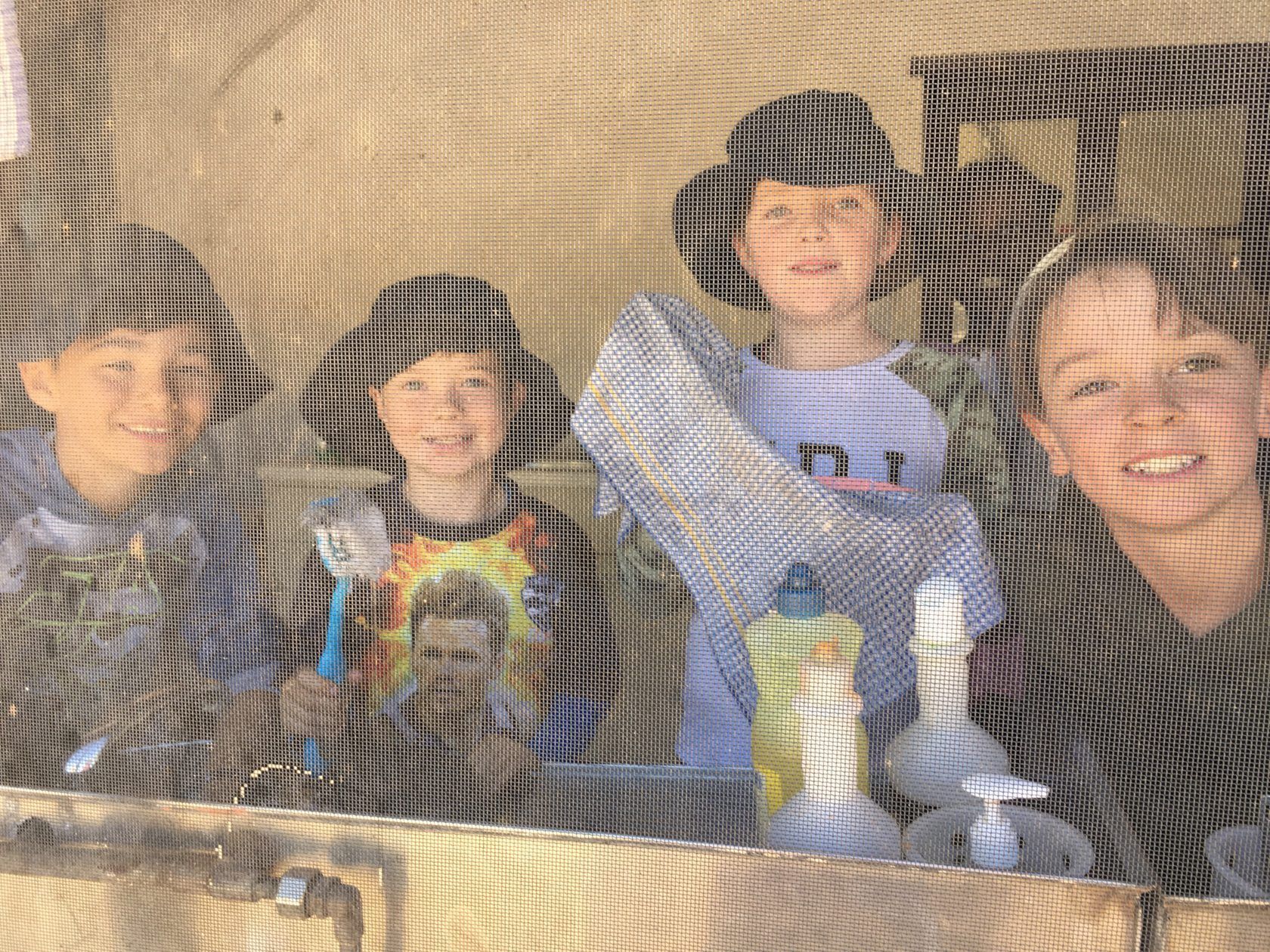 Four children smiling behind a screen, holding cleaning supplies and posing outside.