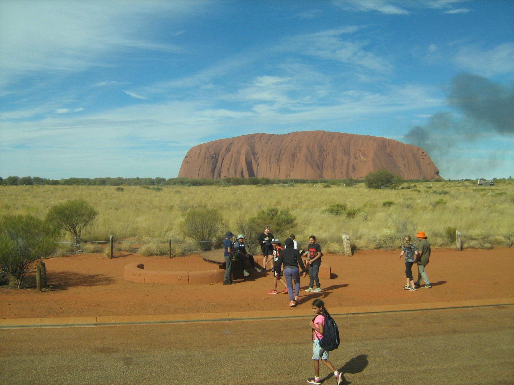 People at Uluru, Australia, standing on red earth, with the large red rock formation and blue sky in the background.