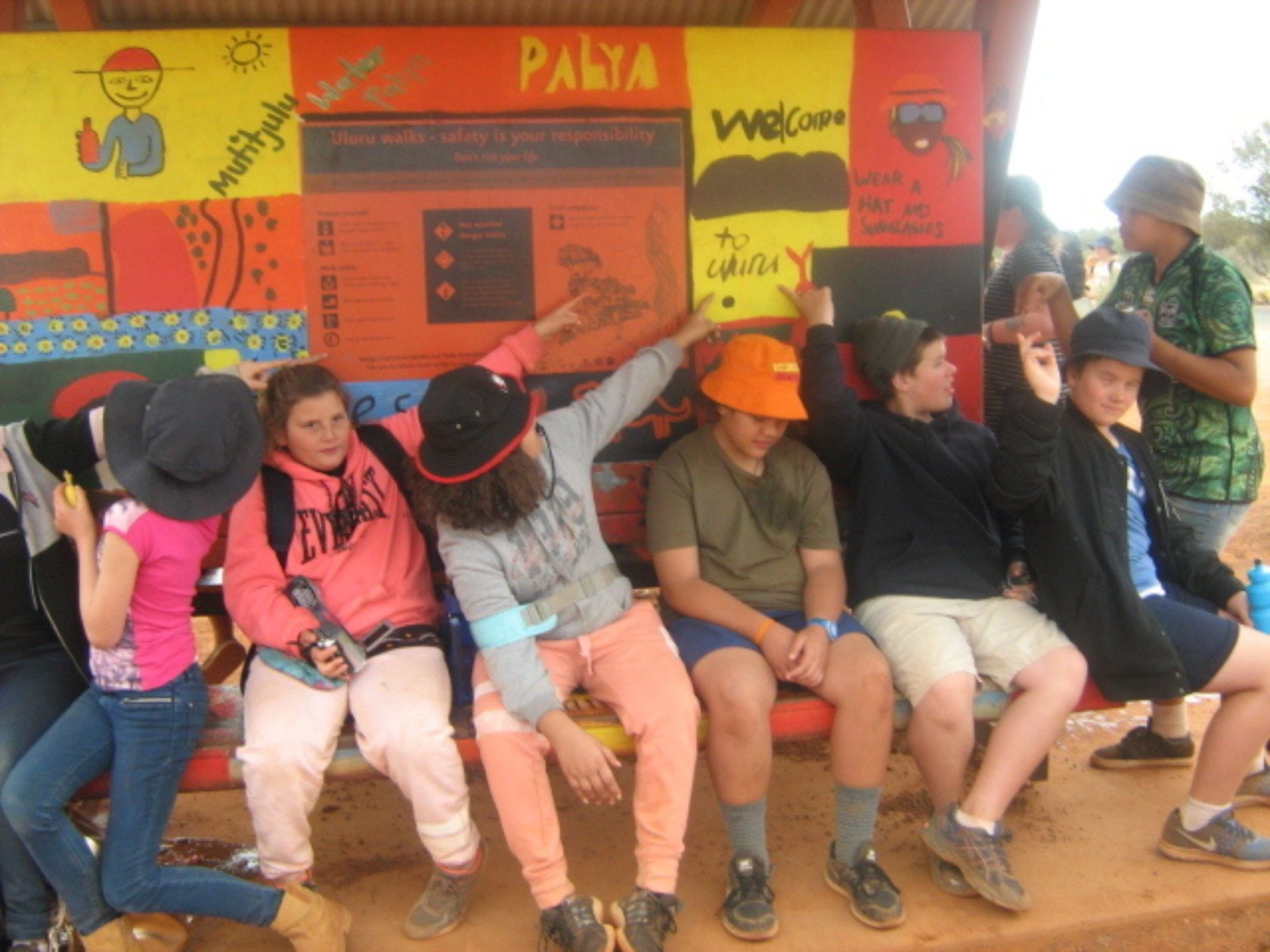 Group of children pose on a painted bench outdoors; some pointing, bright colors in the background.