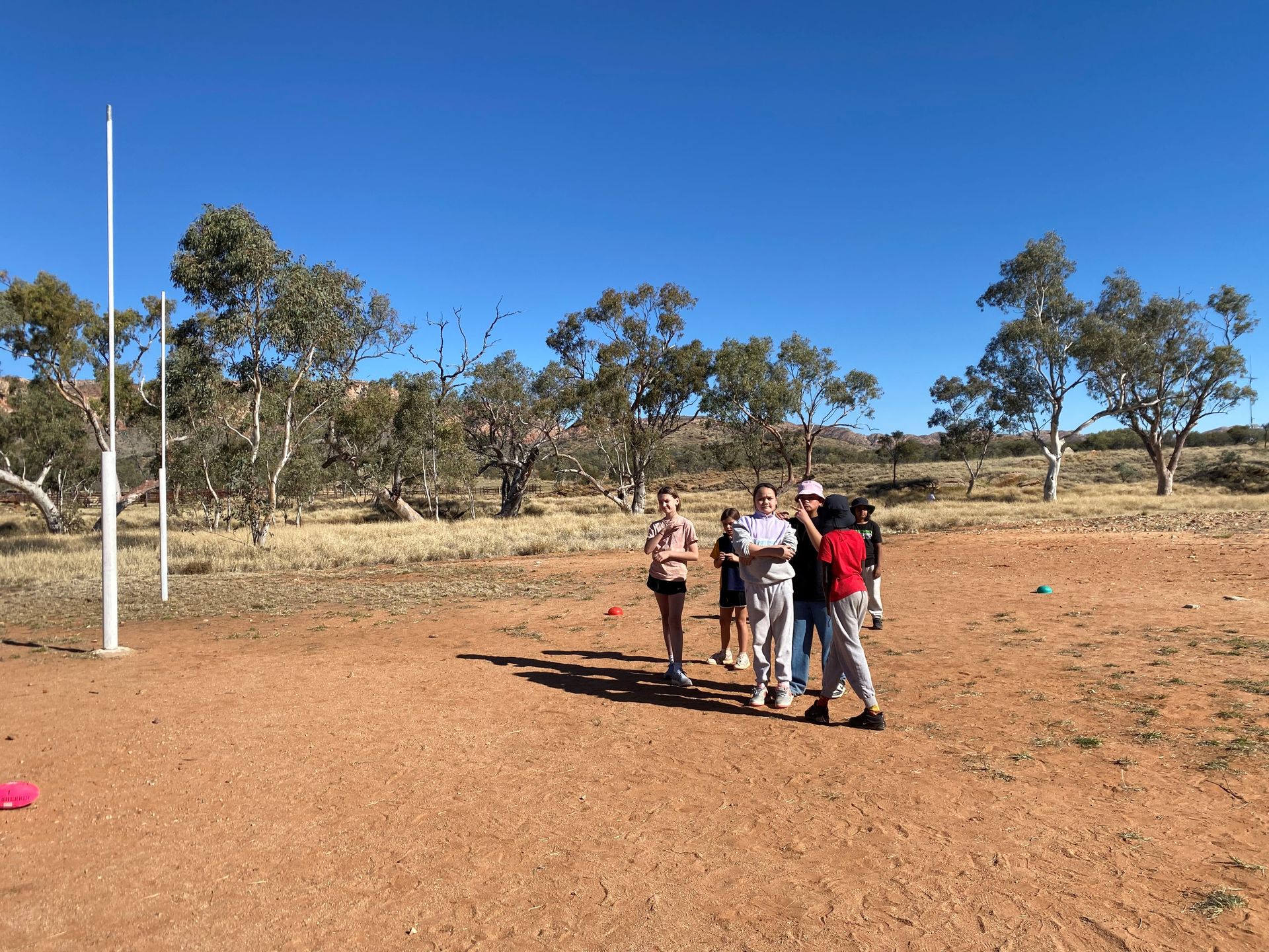 Group of people standing on red dirt field near a goal post, under a bright blue sky.