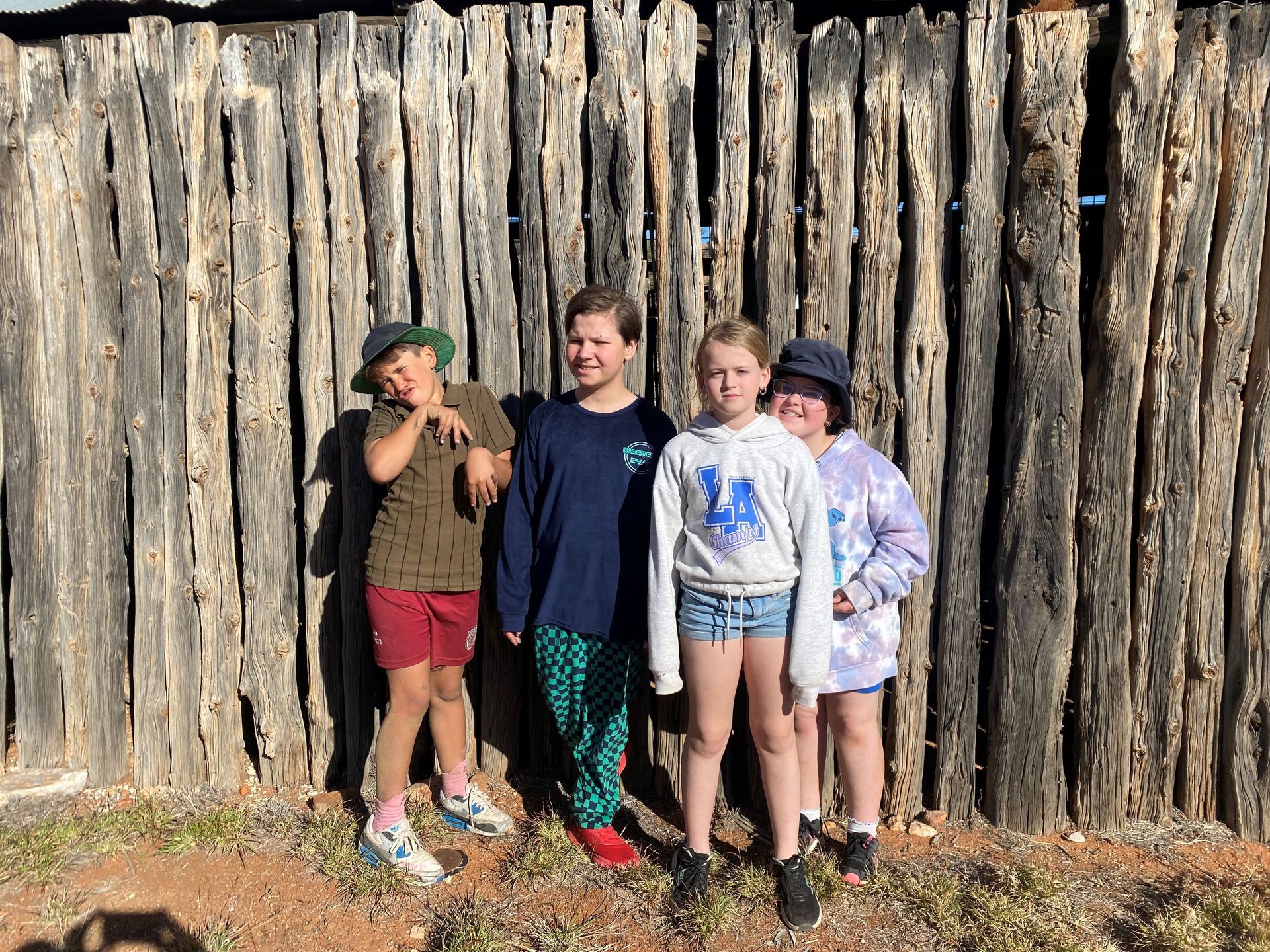 Four kids pose in front of a weathered wooden wall; one makes a silly face.