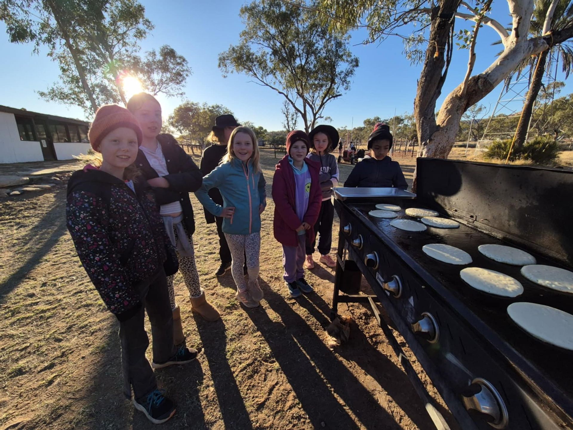 Children watch pancakes cook on a large grill outdoors; sunny day.