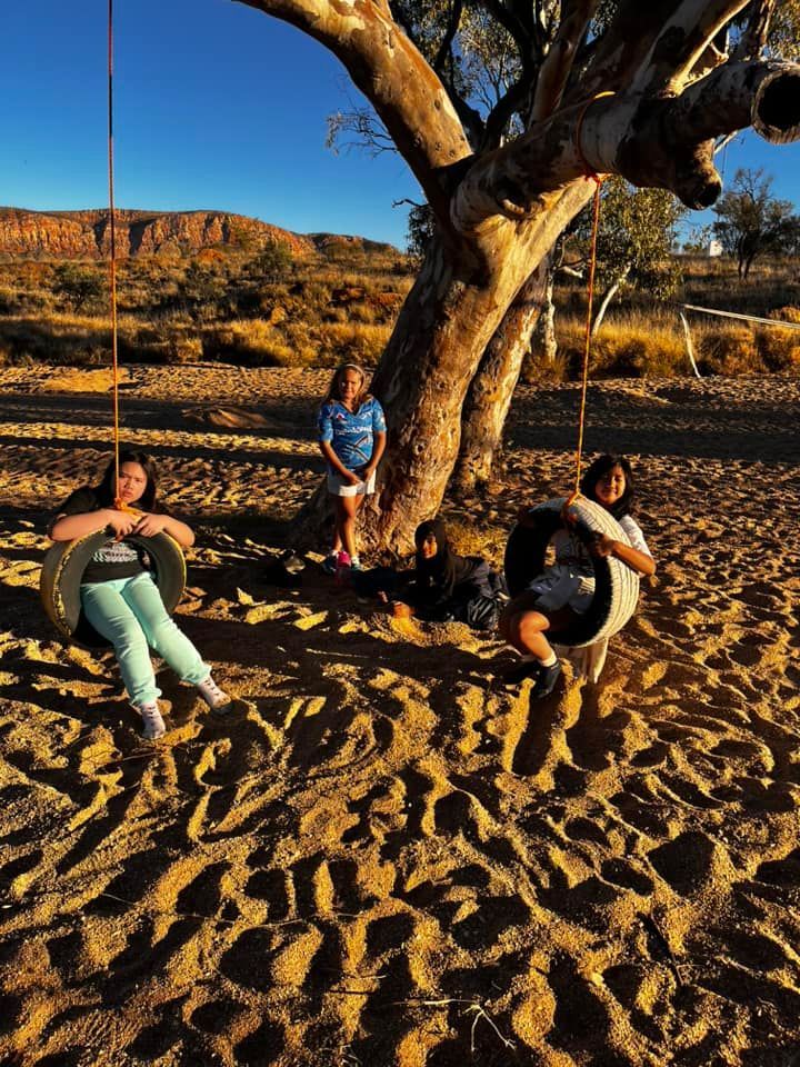 Children on tire swings under a tree in a desert landscape.