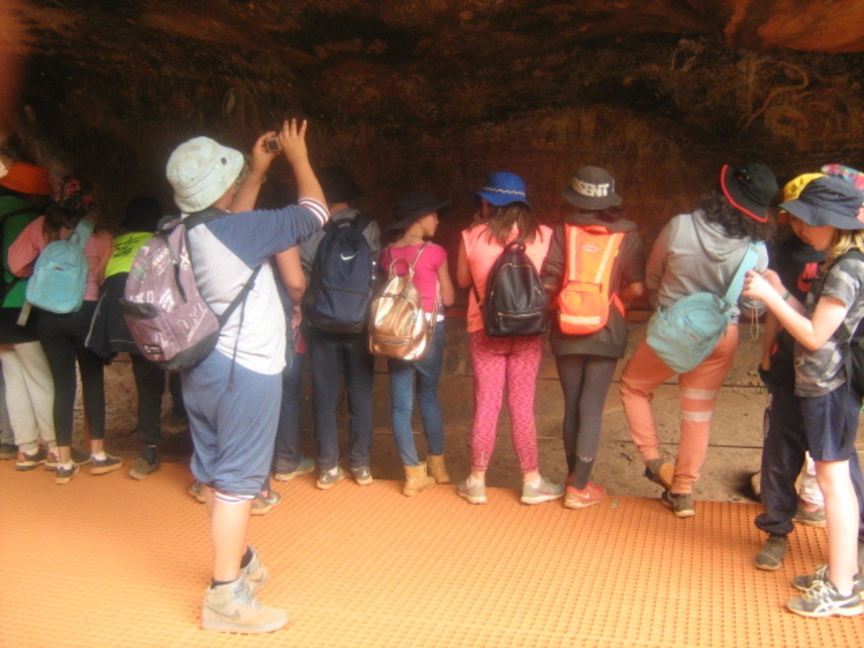 Group of kids looking up at a brown rock ceiling, one taking a photo. Bright clothes, backpacks, inside a cave.