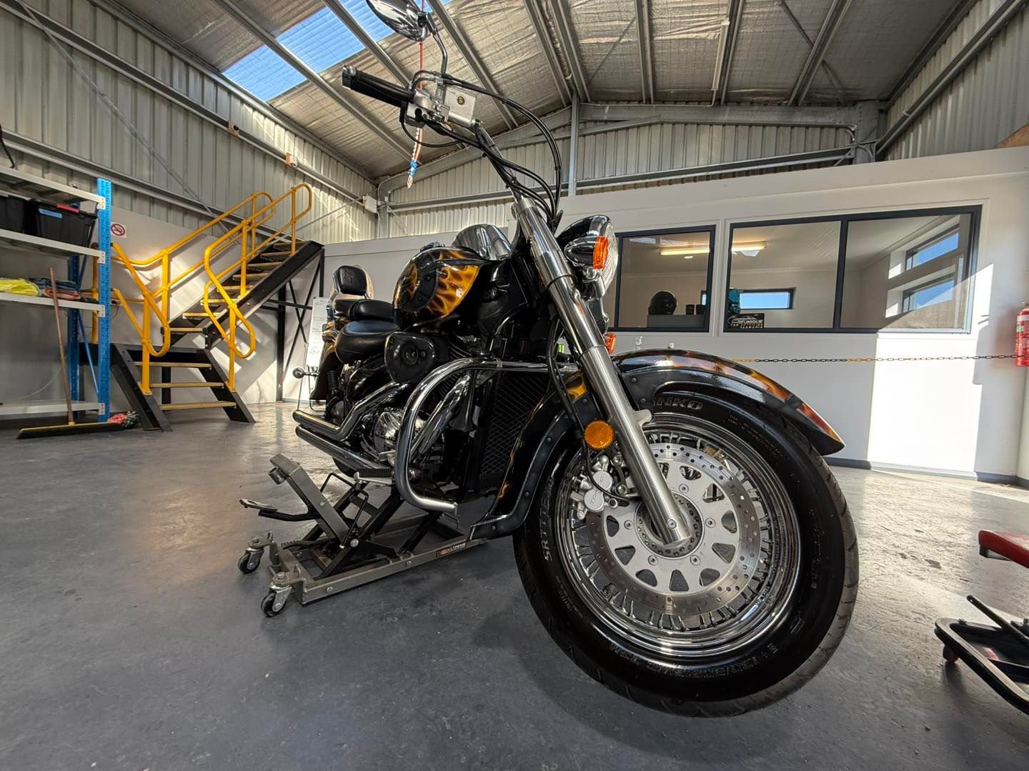Black motorcycle on a lift in a well-lit garage with a yellow staircase and office in the background.
