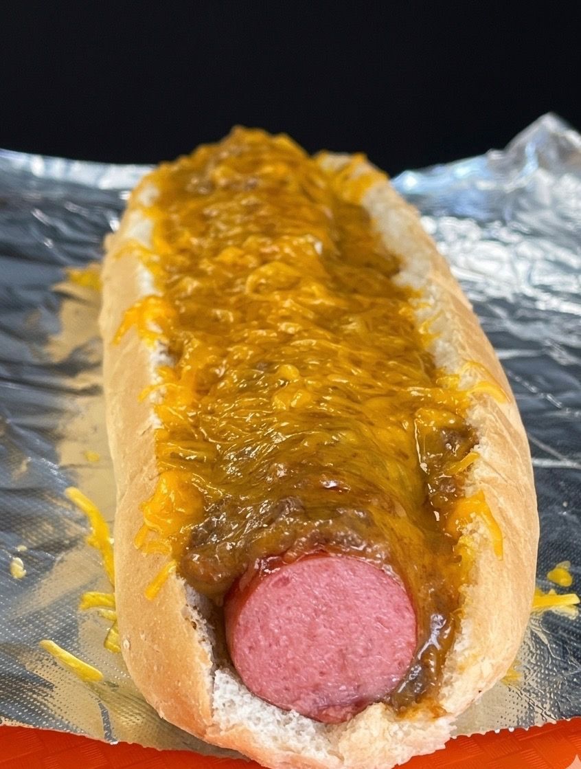 A chili cheese dog in a paper food tray sits on an orange surface.