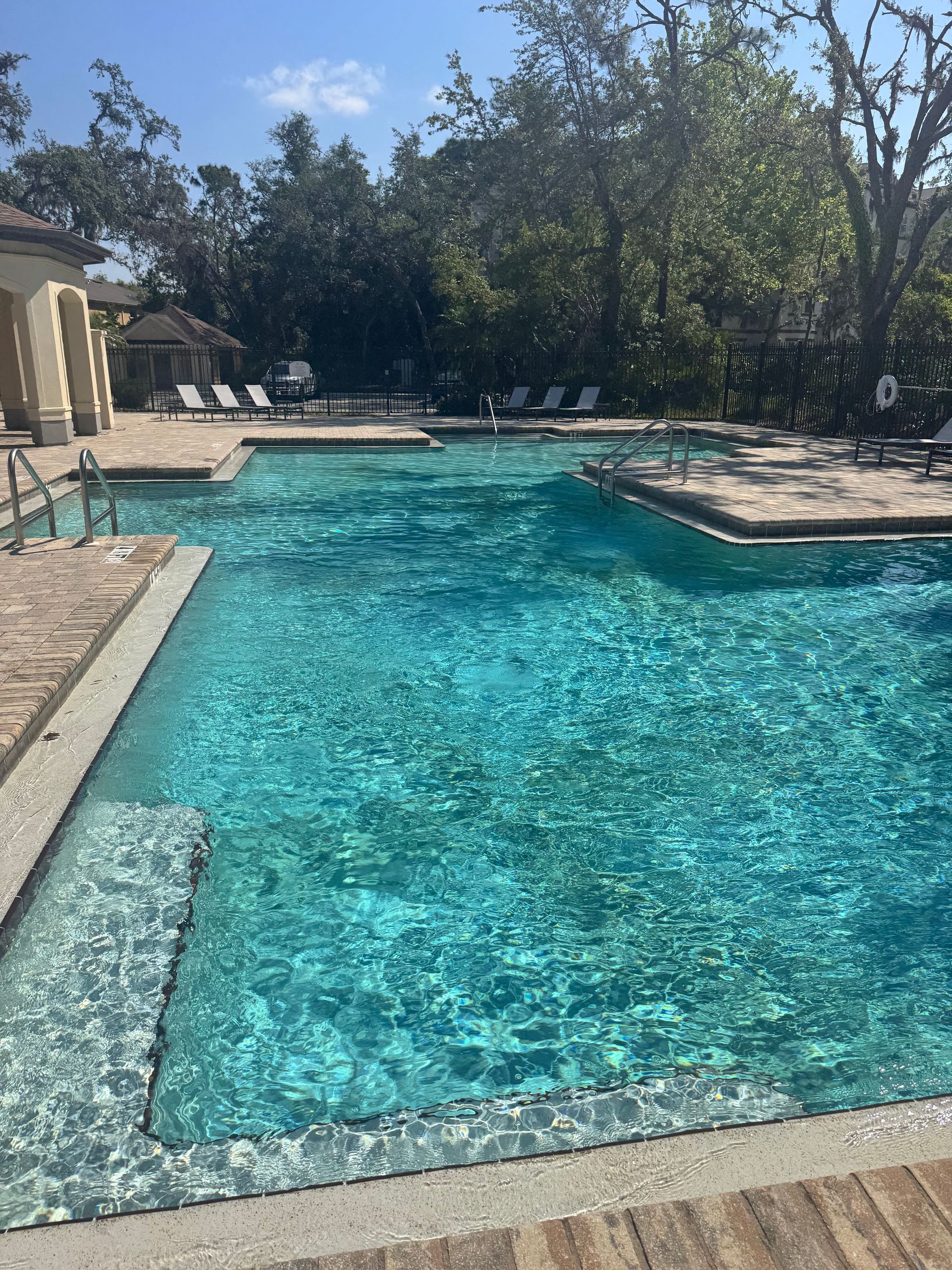 Swimming pool with clear, turquoise water surrounded by stone decking and trees under a bright blue sky.