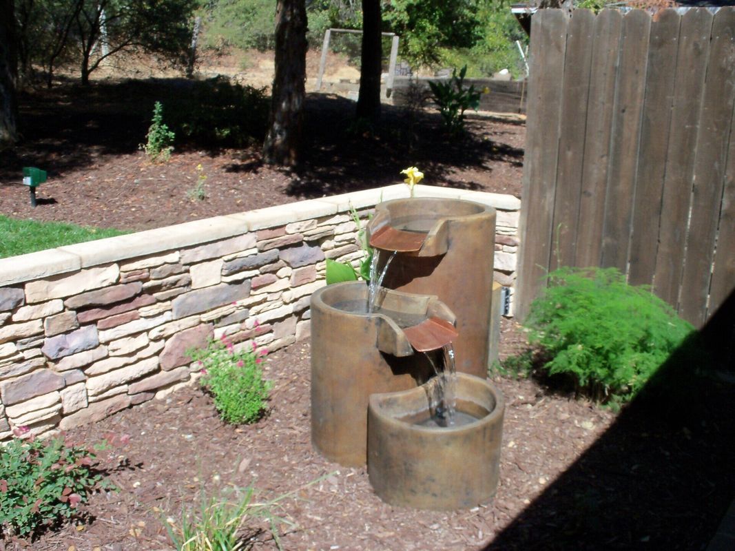 Stone water fountain in a landscaped yard, with tiered cylinders and water flowing.