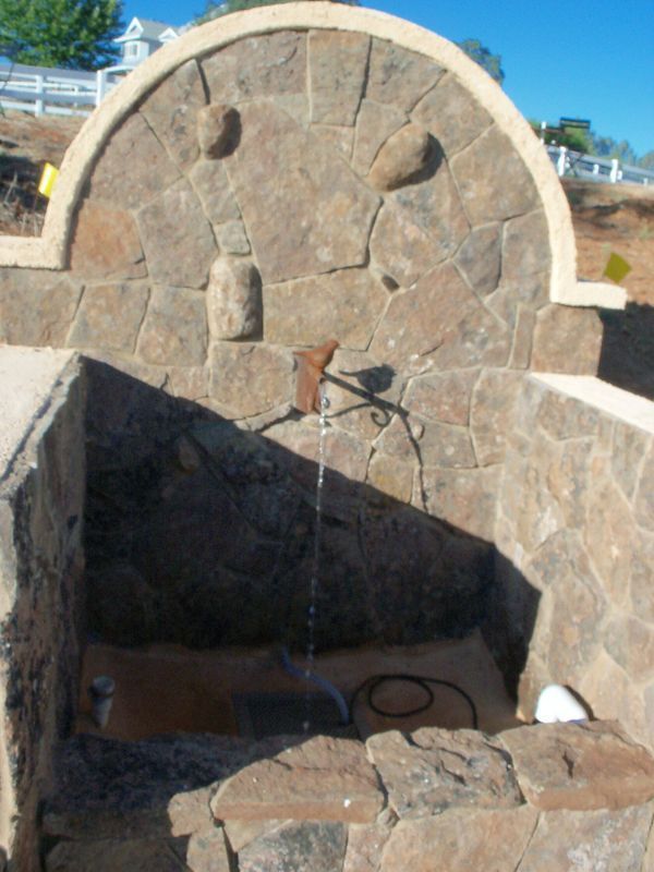 Stone water fountain with arched top and spout, water flowing into basin, outdoor setting.