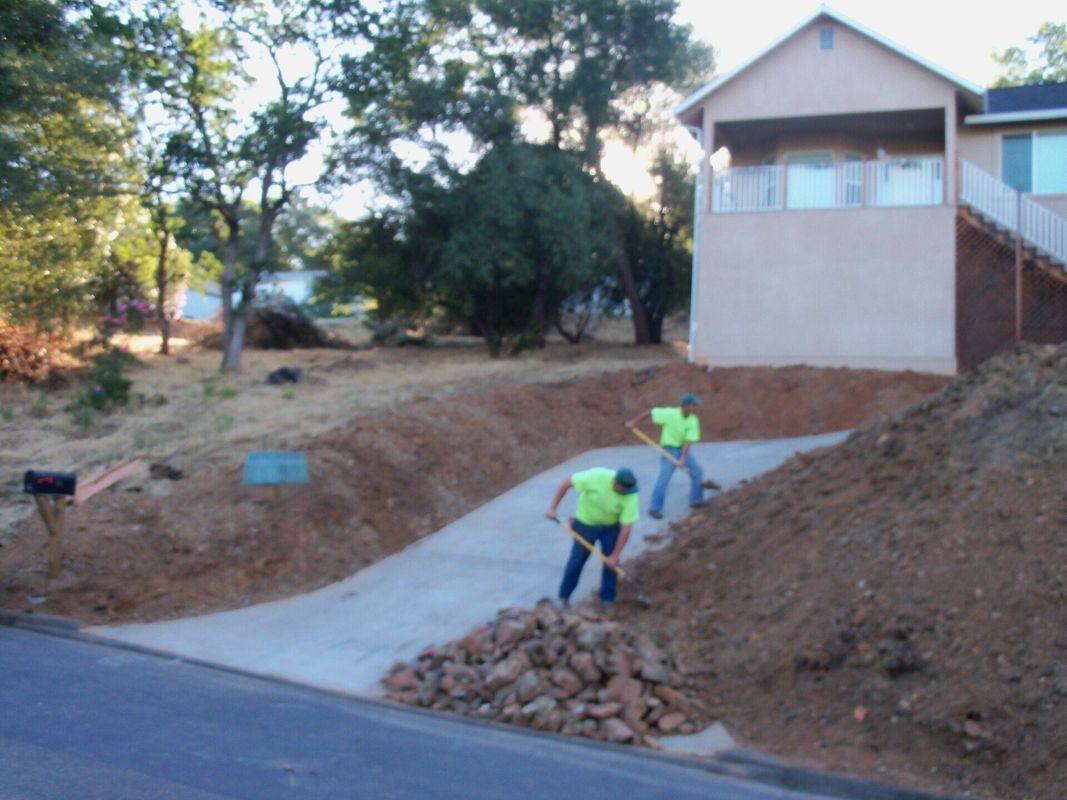 Two workers shoveling dirt alongside a concrete driveway near a house.