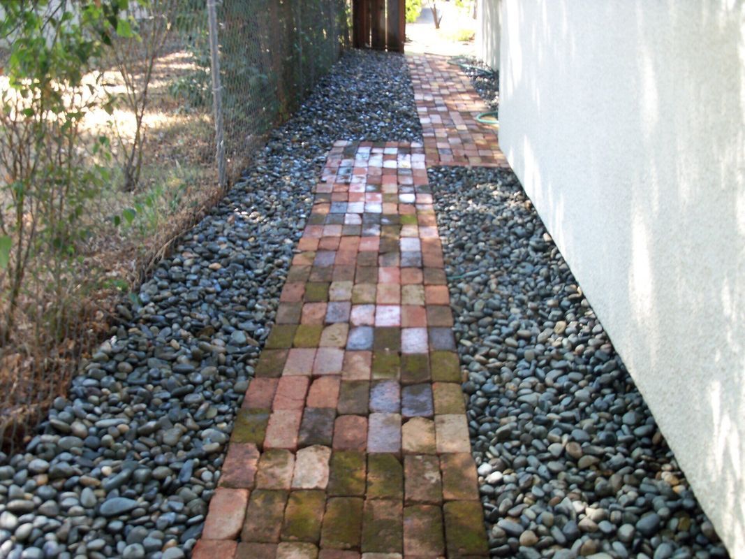 Brick path flanked by gray stones, alongside a white wall and chain-link fence.