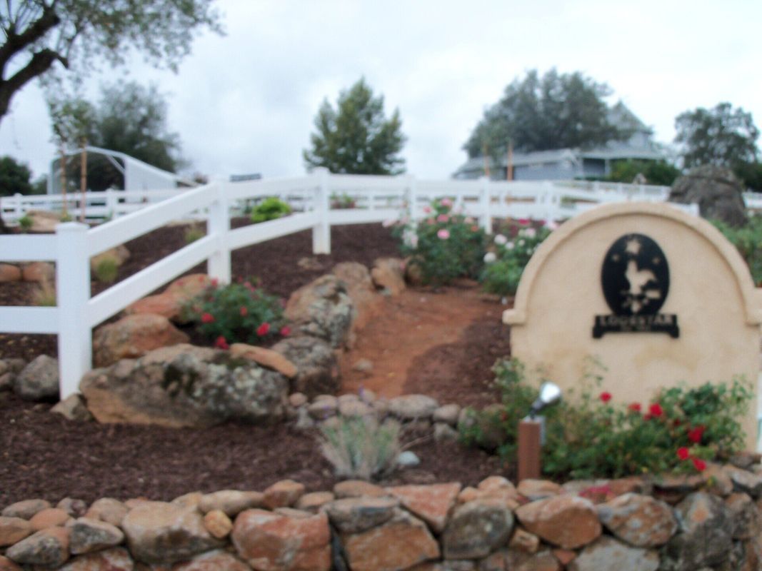 Stone and wood entry sign with a horse graphic, flower garden, and white fence.
