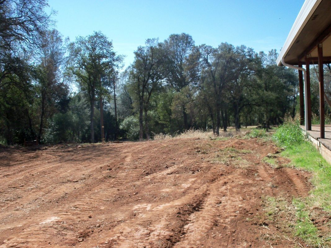 Cleared, uneven dirt area next to a building, with trees in the background under a blue sky.