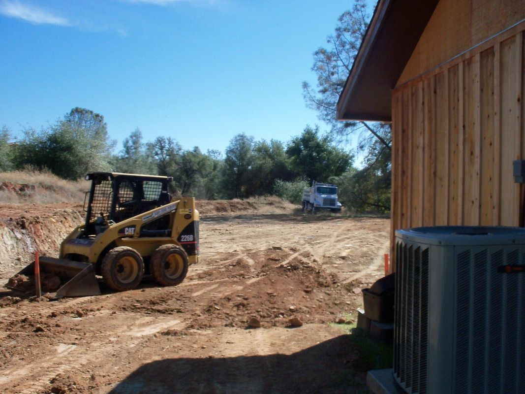 A yellow skid steer on a construction site next to a building; a white truck is in the background.