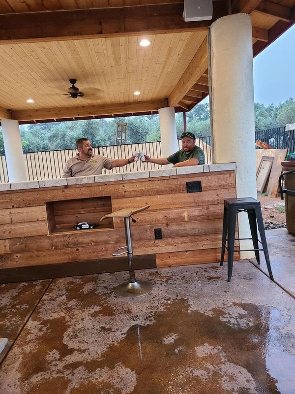 Two men toasting drinks at an outdoor bar with wooden counter under a covered area.
