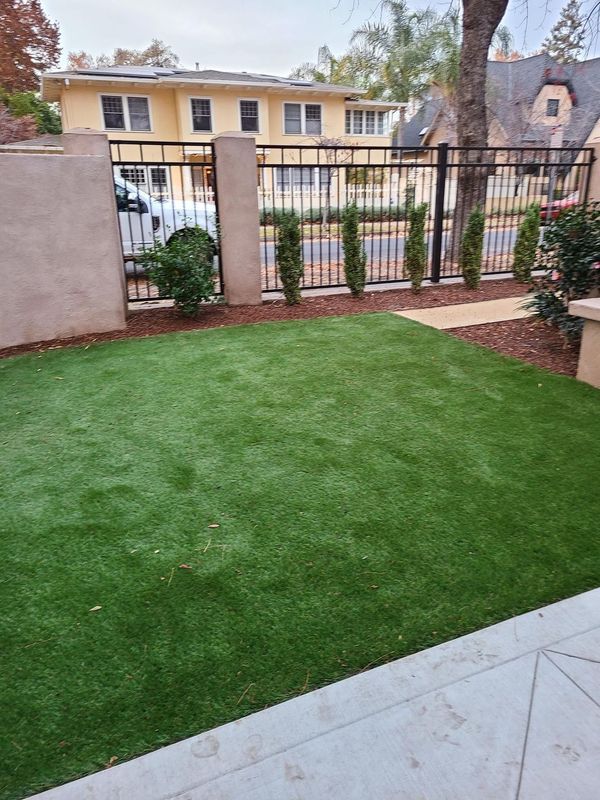 A small yard with artificial green turf, shrubs, and a black fence in front of a beige building.