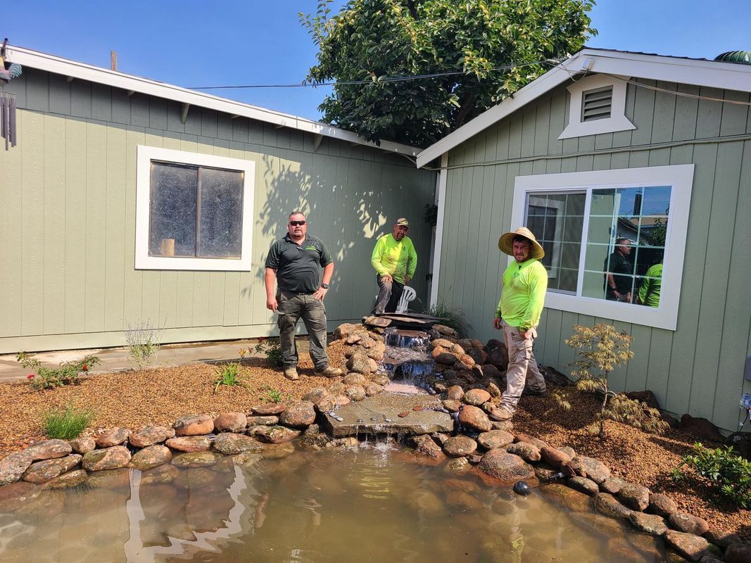 Three people stand near a small pond and waterfall in front of two houses.