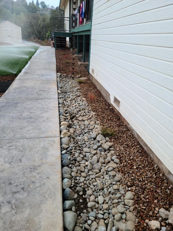 Concrete sidewalk next to a rock-filled drainage channel alongside a white-sided building. Sprinkler waters lawn.