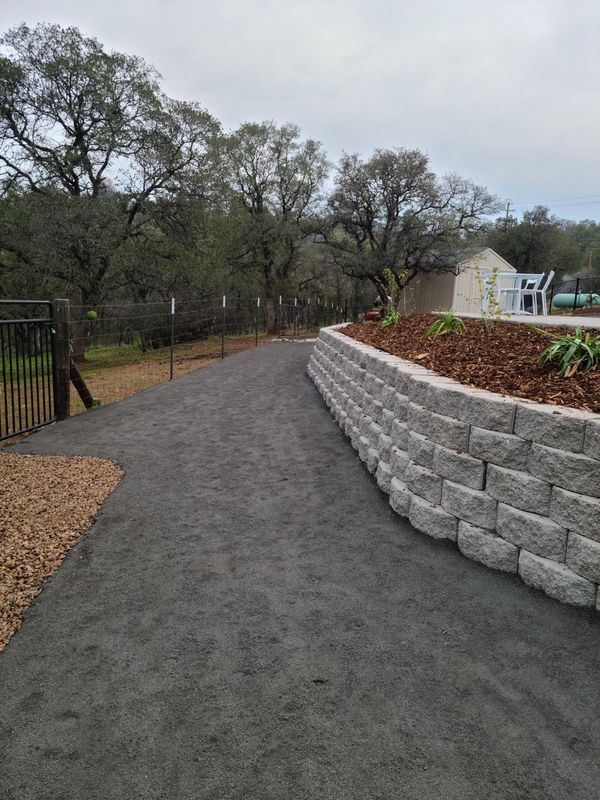 Gravel pathway next to a stacked stone retaining wall. Trees and a building are in the background.