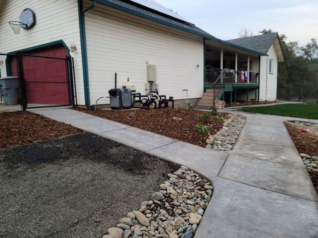 A concrete path curves near a house with a red garage door. Gravel and mulch border the path.