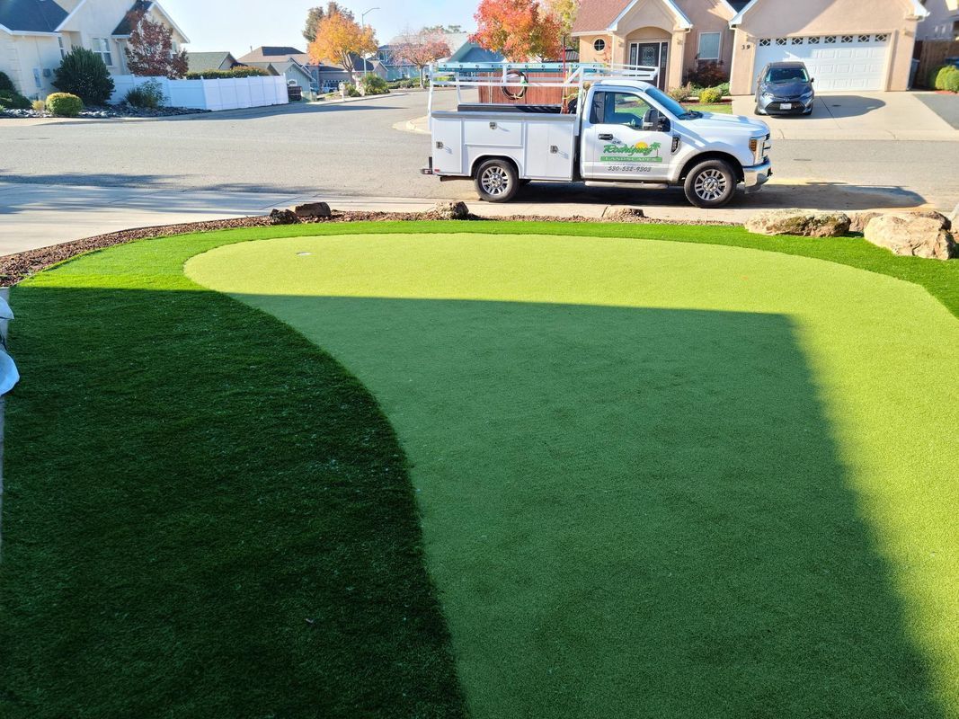 Artificial putting green in a yard with a white work truck parked nearby. Sunny day.