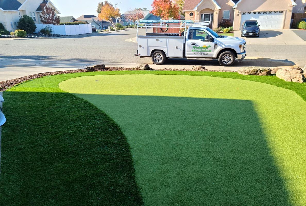 Green artificial turf golf putting green in a front yard; white truck parked in the street.