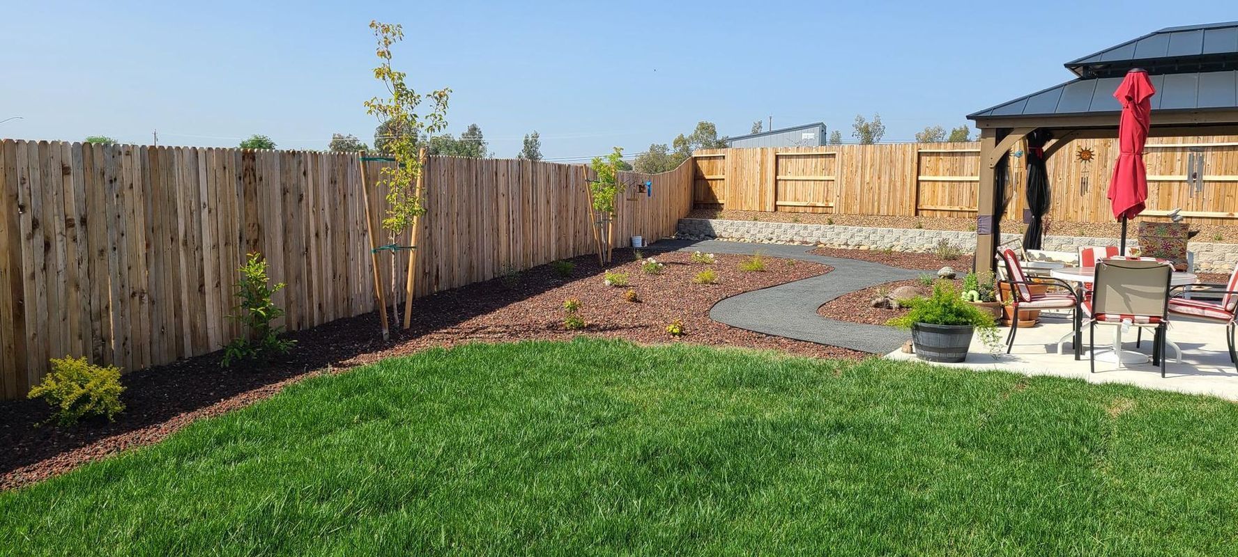 A backyard with green grass, a wooden fence, a stone pathway, a gazebo, and a red umbrella.