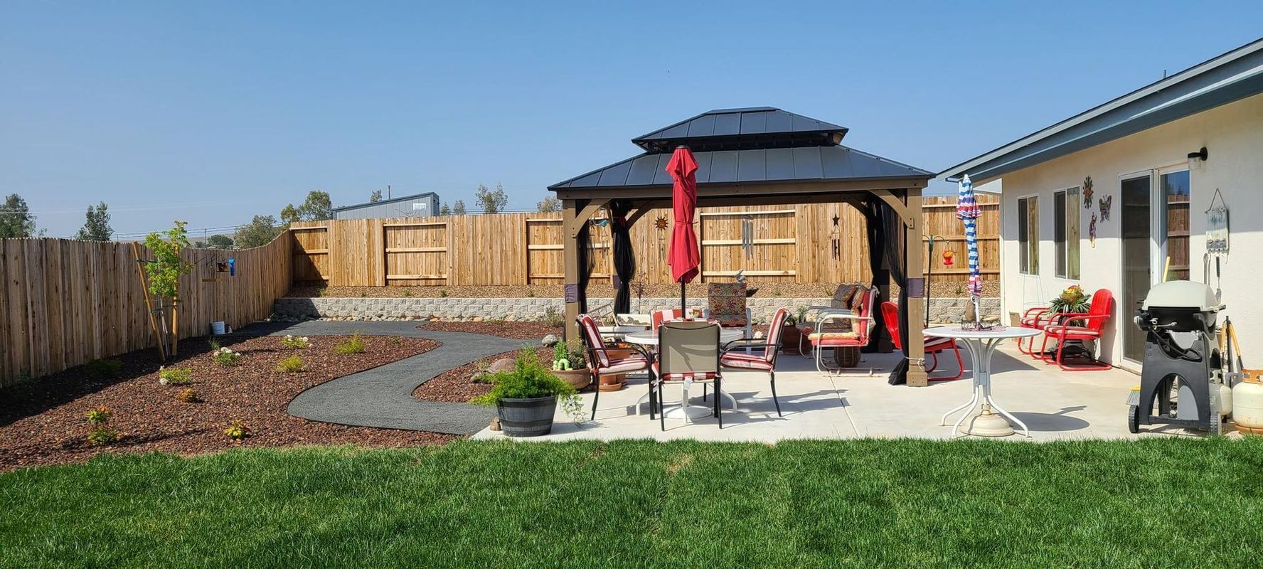 Backyard with gazebo, patio furniture, pathway, and lush green lawn under a clear blue sky.