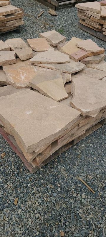 Stone pavers stacked on a wooden pallet, with other piles of pavers in the background on gravel.