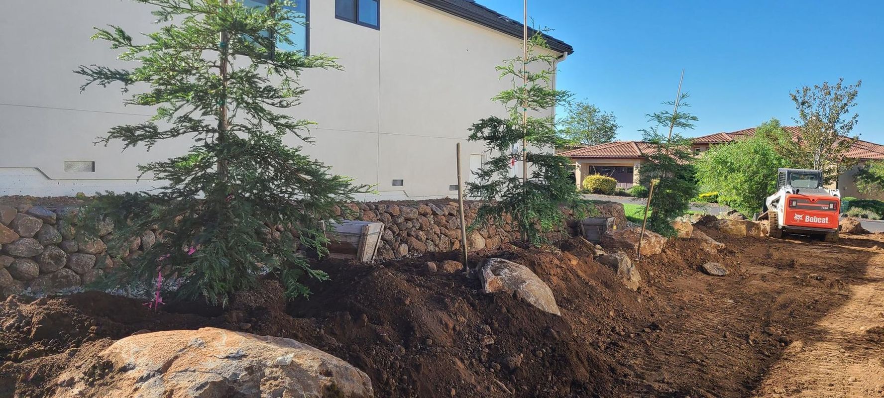 Construction site near a house, with a small excavator, trees, and fresh soil. Clear, blue sky.