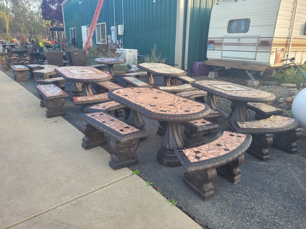 Outdoor display of mosaic-tiled picnic tables and benches on a concrete surface, near a green building and trailer.