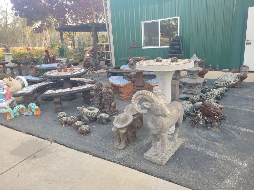 Garden statues and furniture displayed outside a green building: ram statue, bird baths, tables, and sculptures.