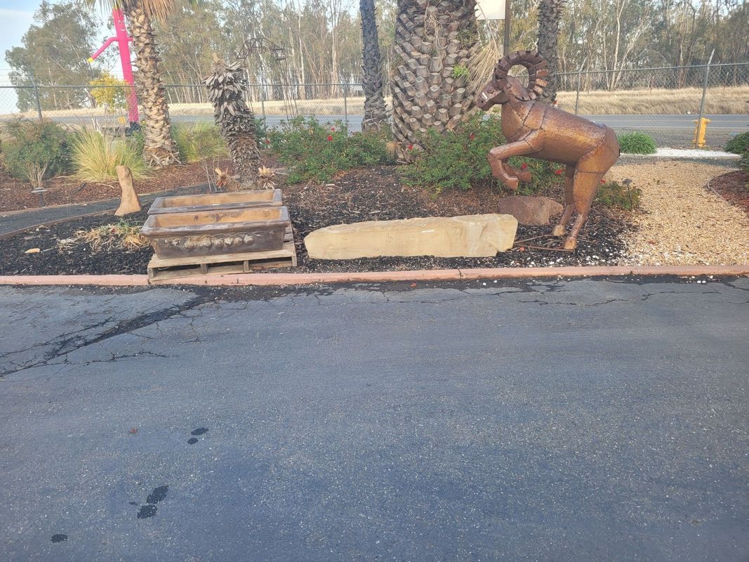 Bronze ram statue beside a landscaped garden with trees, on the edge of a paved area.