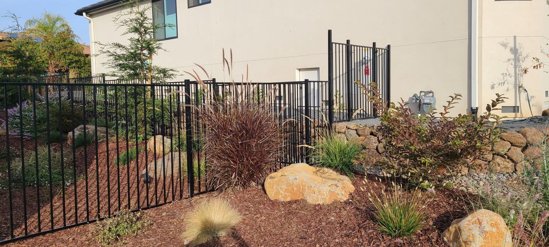Black metal fence with landscaping, rocks, and a beige house in the background.