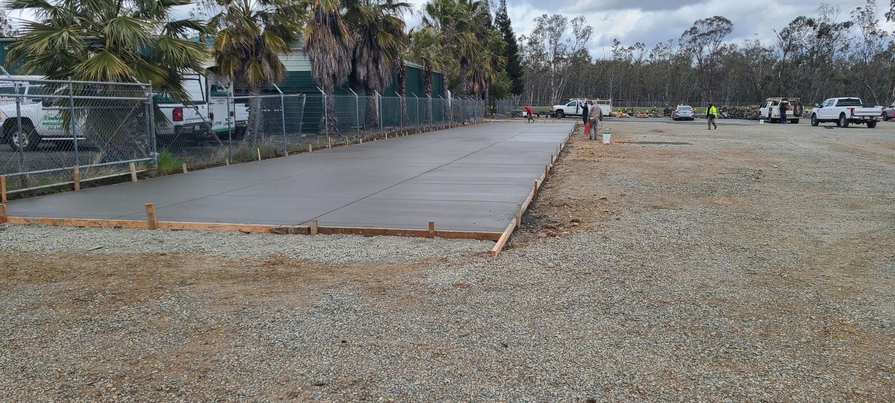 Freshly poured concrete slab next to a gravel parking area with several white work trucks and a fence in the background.